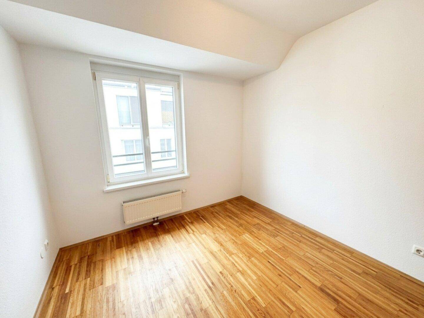 Helles Schlafzimmer mit Holzboden, großem Fenster und Heizkörper, bereit zur individuellen Gestaltung.
