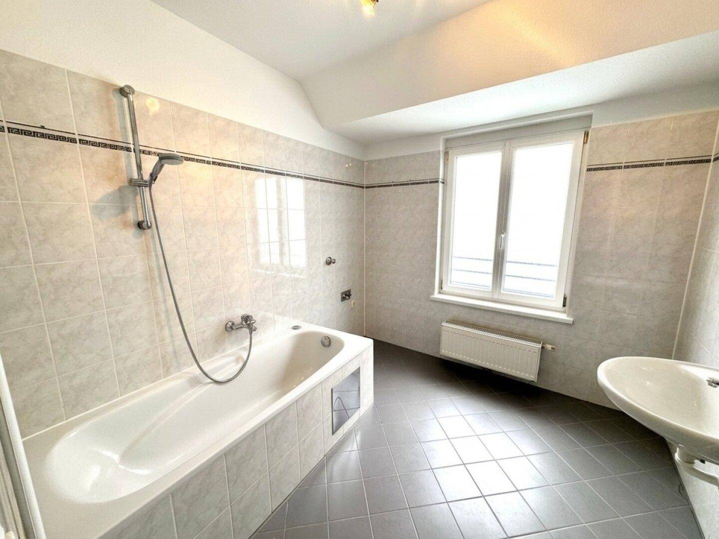 Helles Badezimmer mit Badewanne, Waschbecken und Fenster, komplett gefliest.