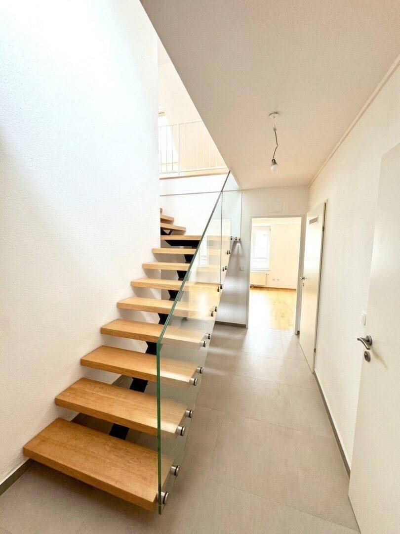 Heller Flur mit moderner Holztreppe und Glasgeländer, führt zu den oberen Etagen.