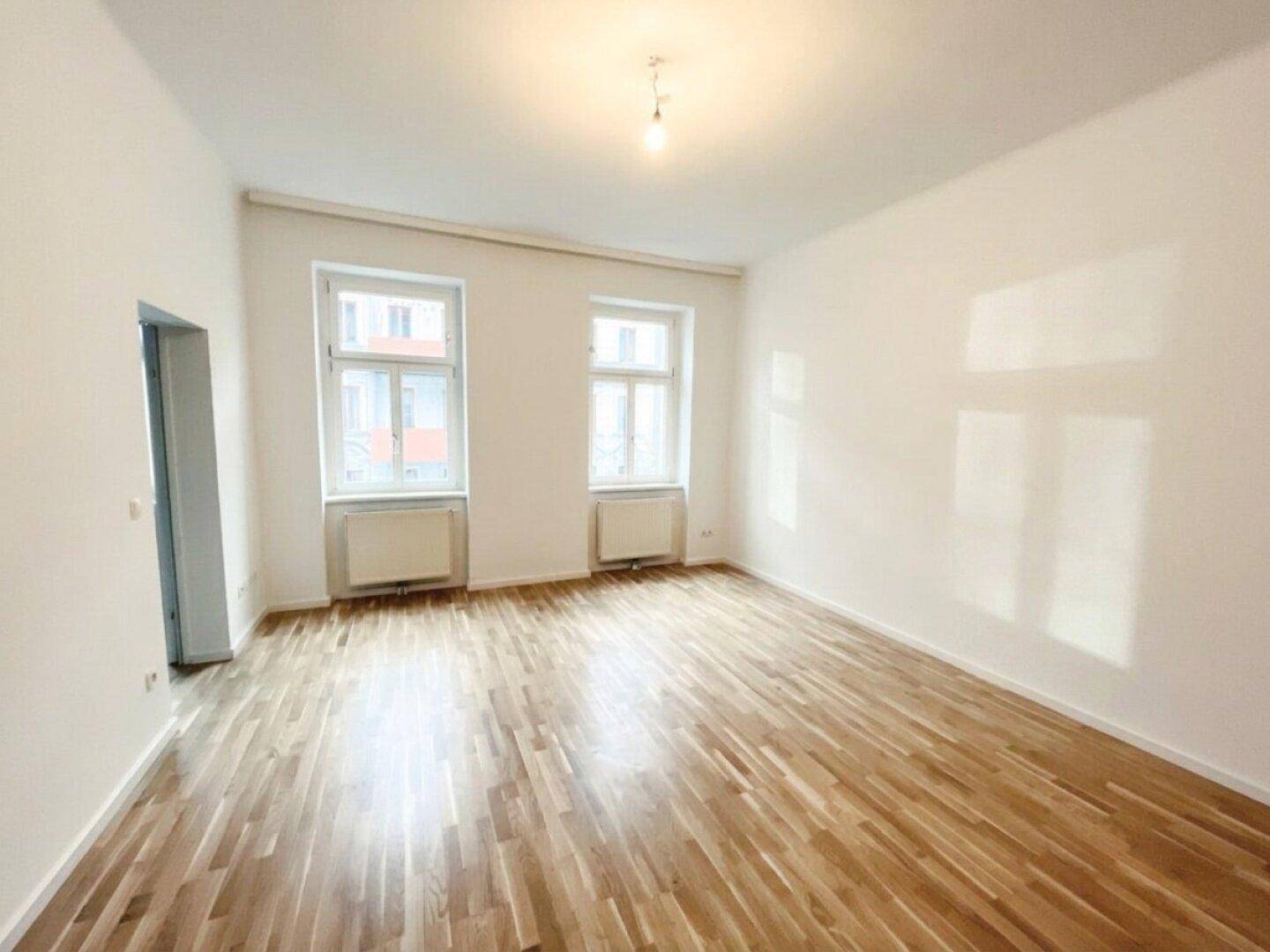 Geräumiges, helles Zimmer mit Holzboden und großen Fenstern, ideal für Wohnzwecke.
