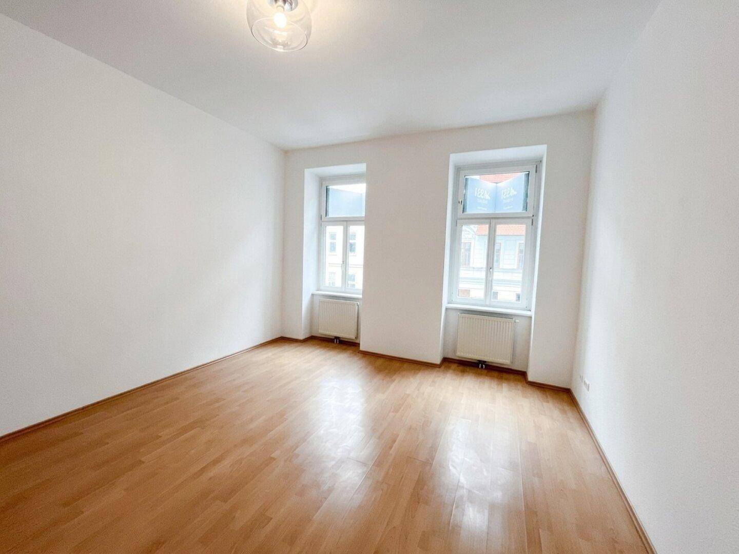 Freundliches Zimmer mit Holzboden und zwei großen Fenstern, ideal für Wohn- oder Schlafbereich.