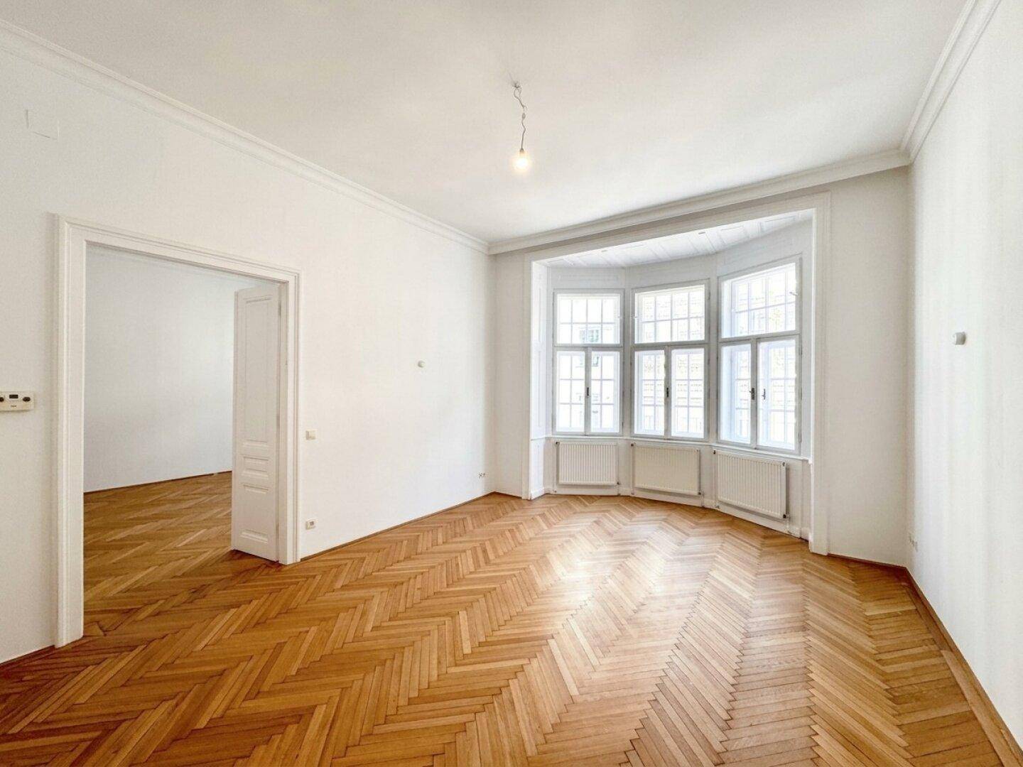 Helles Wohnzimmer mit Fischgrätparkett und einem schönen Erkerfenster, das viel Tageslicht hereinlässt.