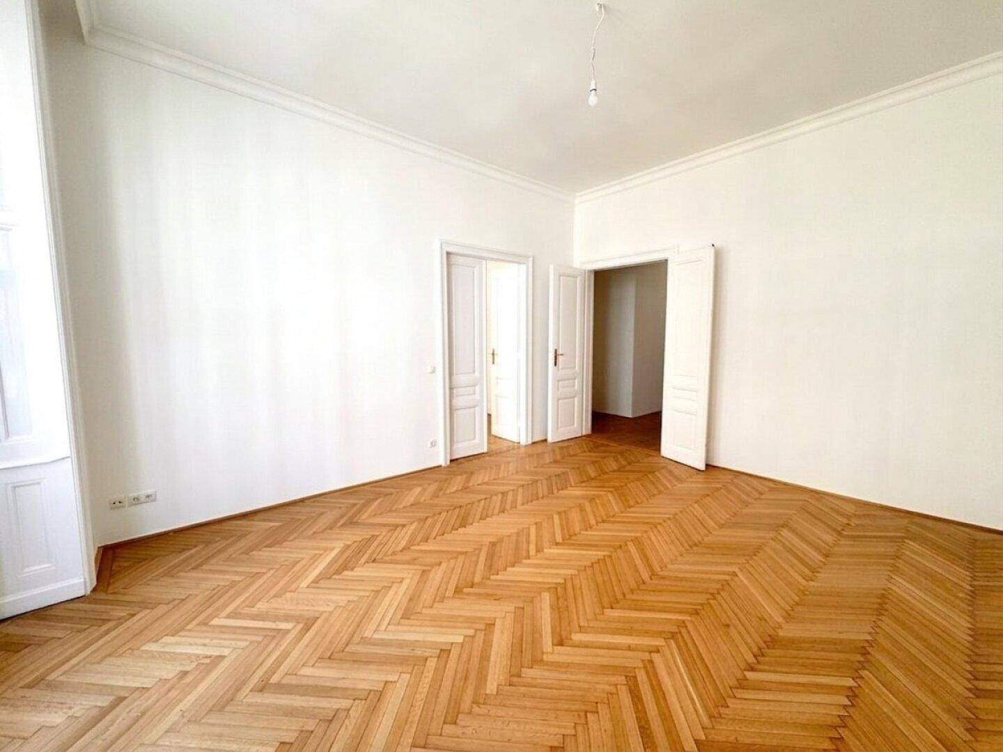 Geräumiges Zimmer mit Fischgrätparkett und zwei Flügeltüren, die zu weiteren Räumen führen.