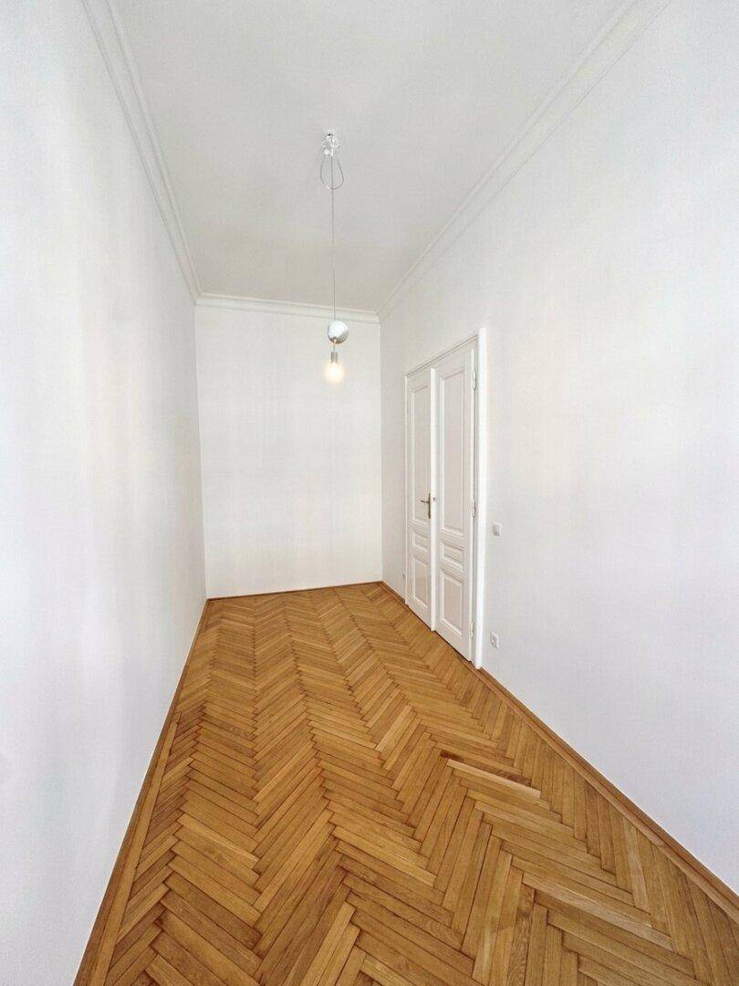 Langer, schmaler Flur mit Fischgrätparkett und weißen Wänden, der zu weiteren Räumen führt.