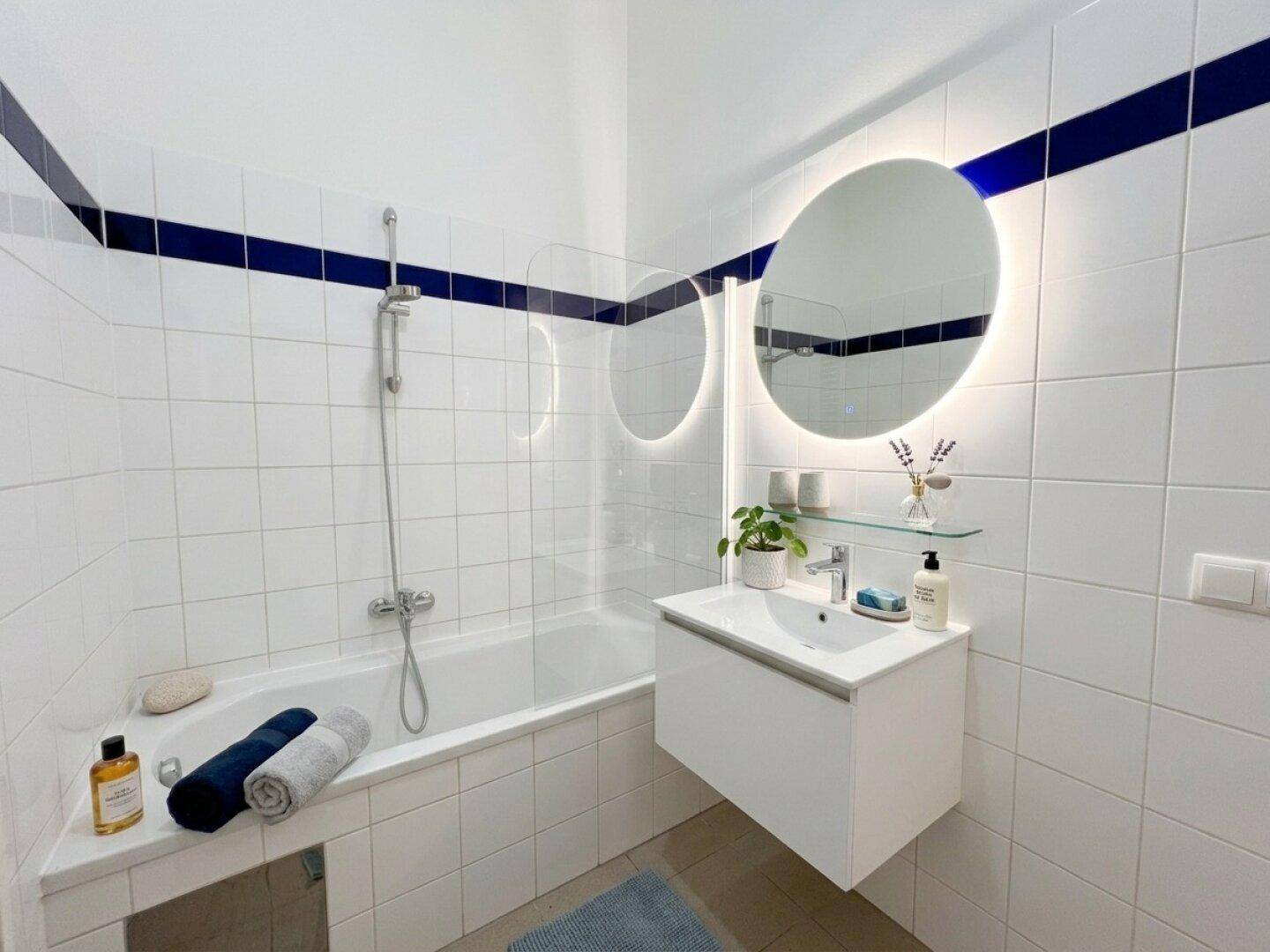 Helles Badezimmer mit Badewanne, modernem Waschbecken und beleuchtetem Spiegel, dekoriert mit Pflanzen.