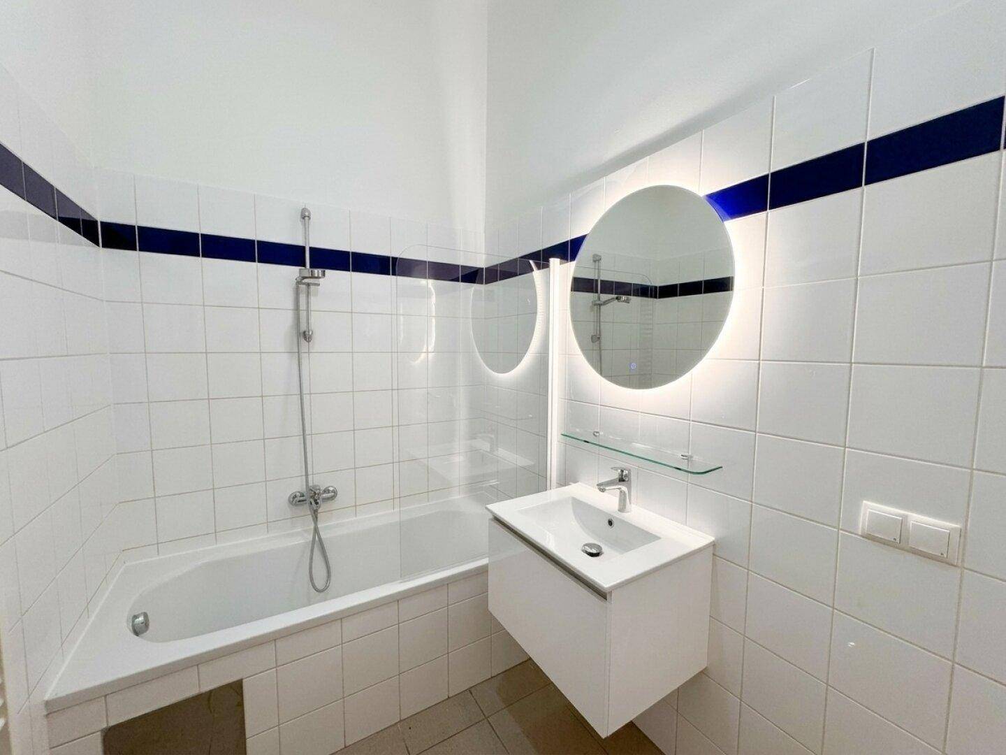 Modernes Badezimmer mit weiß gefliesten Wänden, blauer Akzentlinie und beleuchtetem Spiegel über dem Waschbecken.