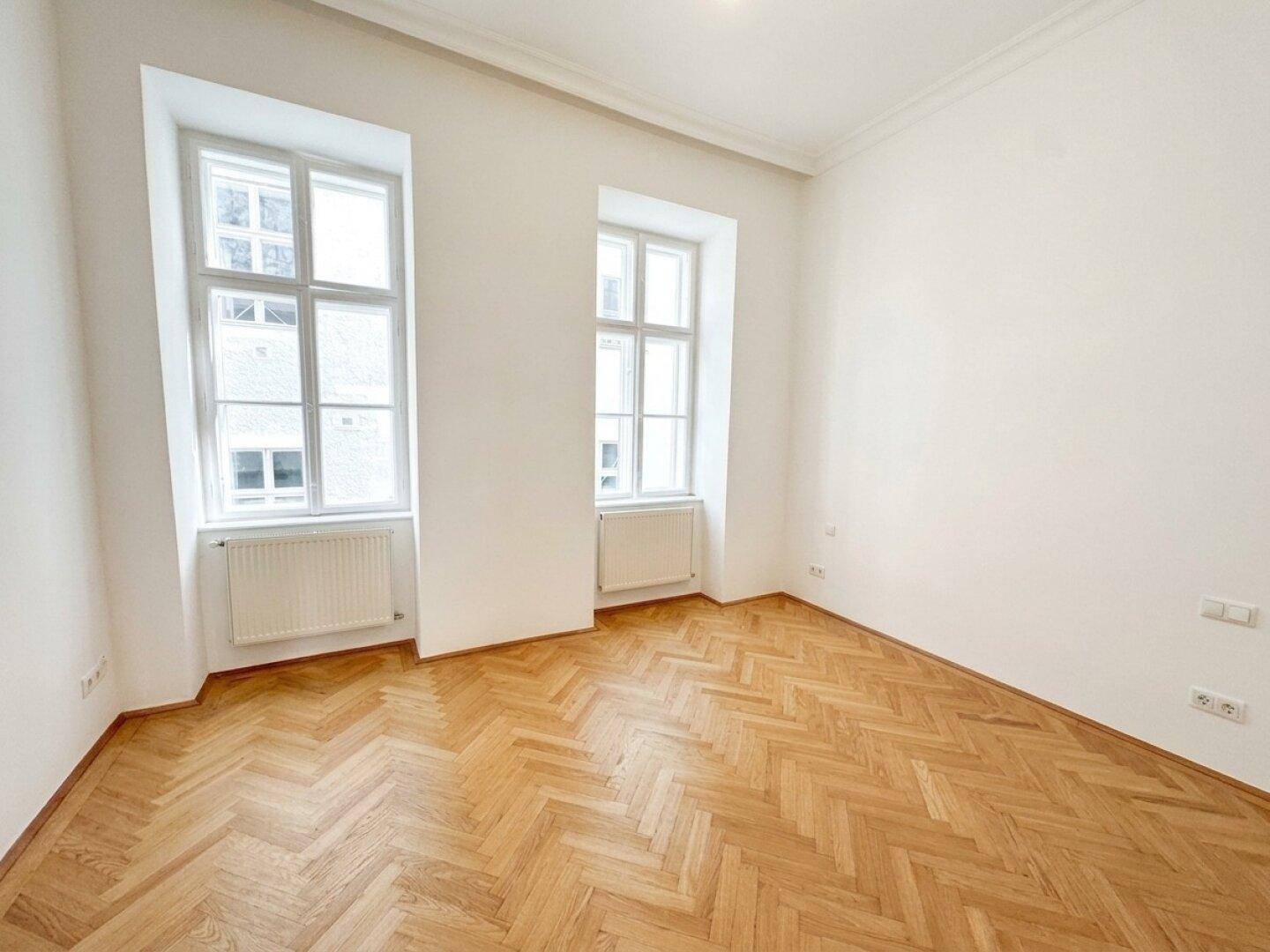 Geräumiges Schlafzimmer mit Fischgrätparkett und zwei hohen Fenstern, die viel Tageslicht bieten.