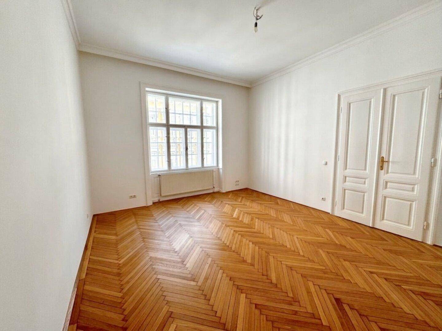 Helles Zimmer mit Fischgrätparkett und einem großen Fenster, das viel Licht in den Raum bringt.