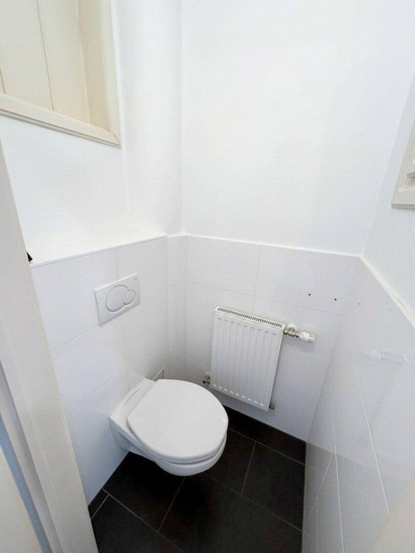 Separates WC mit Wandtoilette und Heizkörper, ausgestattet mit dunklen Bodenfliesen.