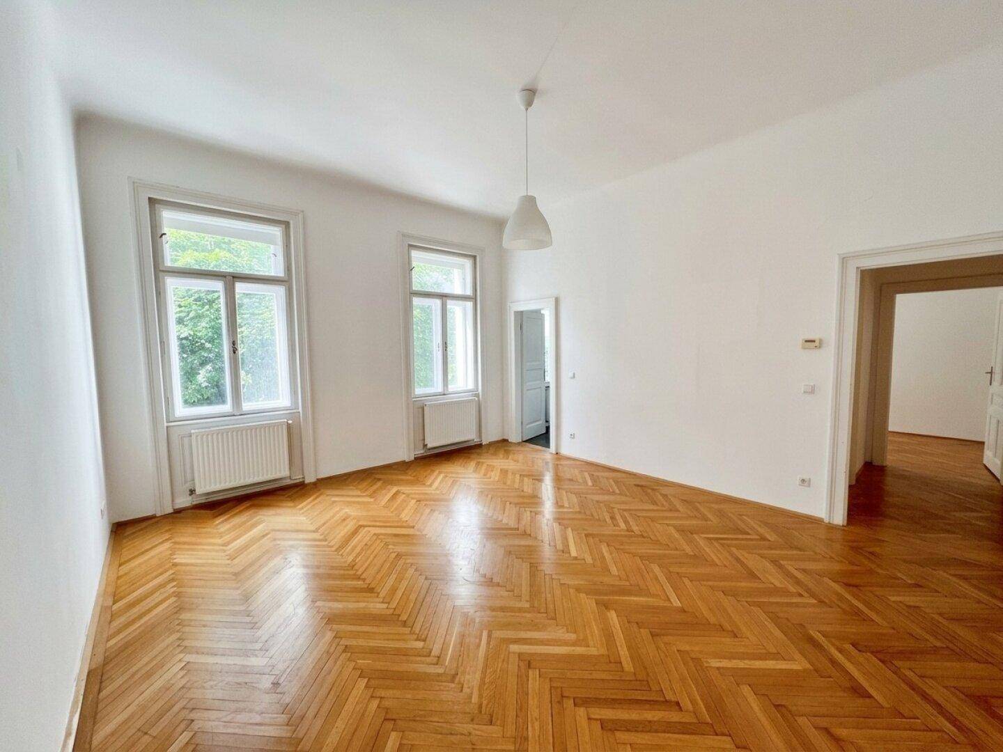 Heller Wohnraum mit elegantem Fischgrätparkett und hohen Fenstern, die viel Licht hereinlassen.
