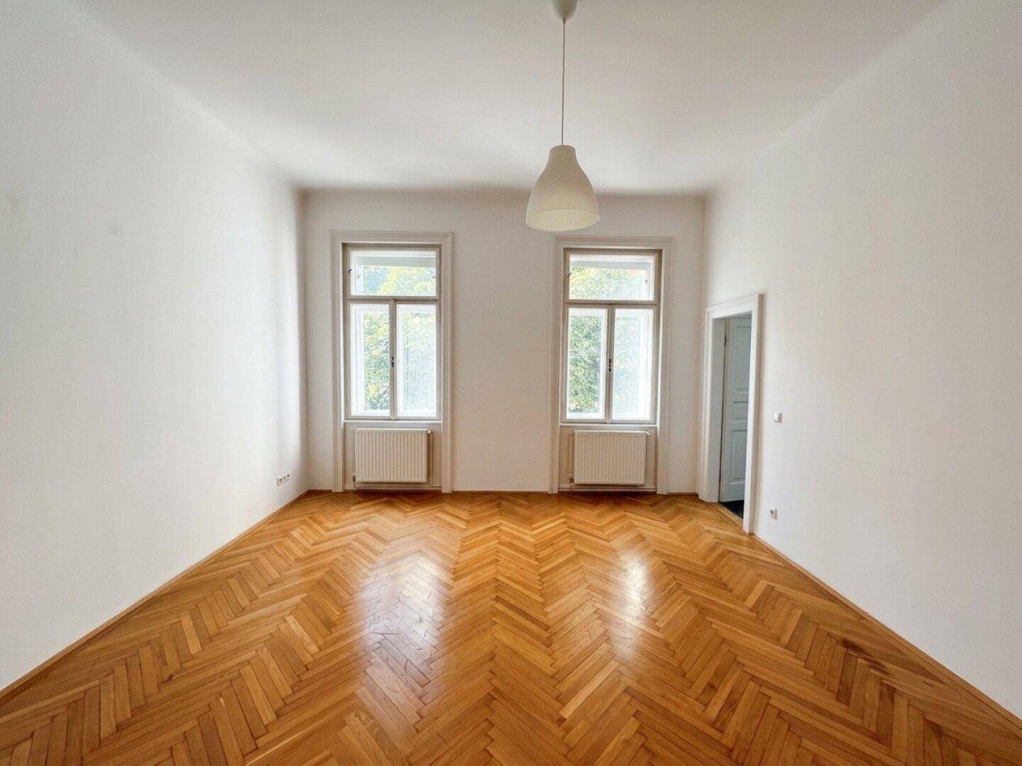 Geräumiges Wohnzimmer mit klassischem Fischgrätparkett und hohen Fenstern, bereit zur individuellen Gestaltung.