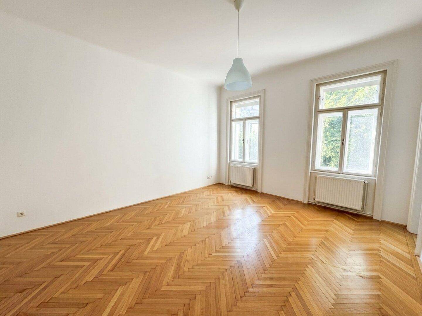 Lichtdurchfluteter Raum mit Fischgrätparkett und zwei großen Fenstern, ideal für ein Wohnzimmer.