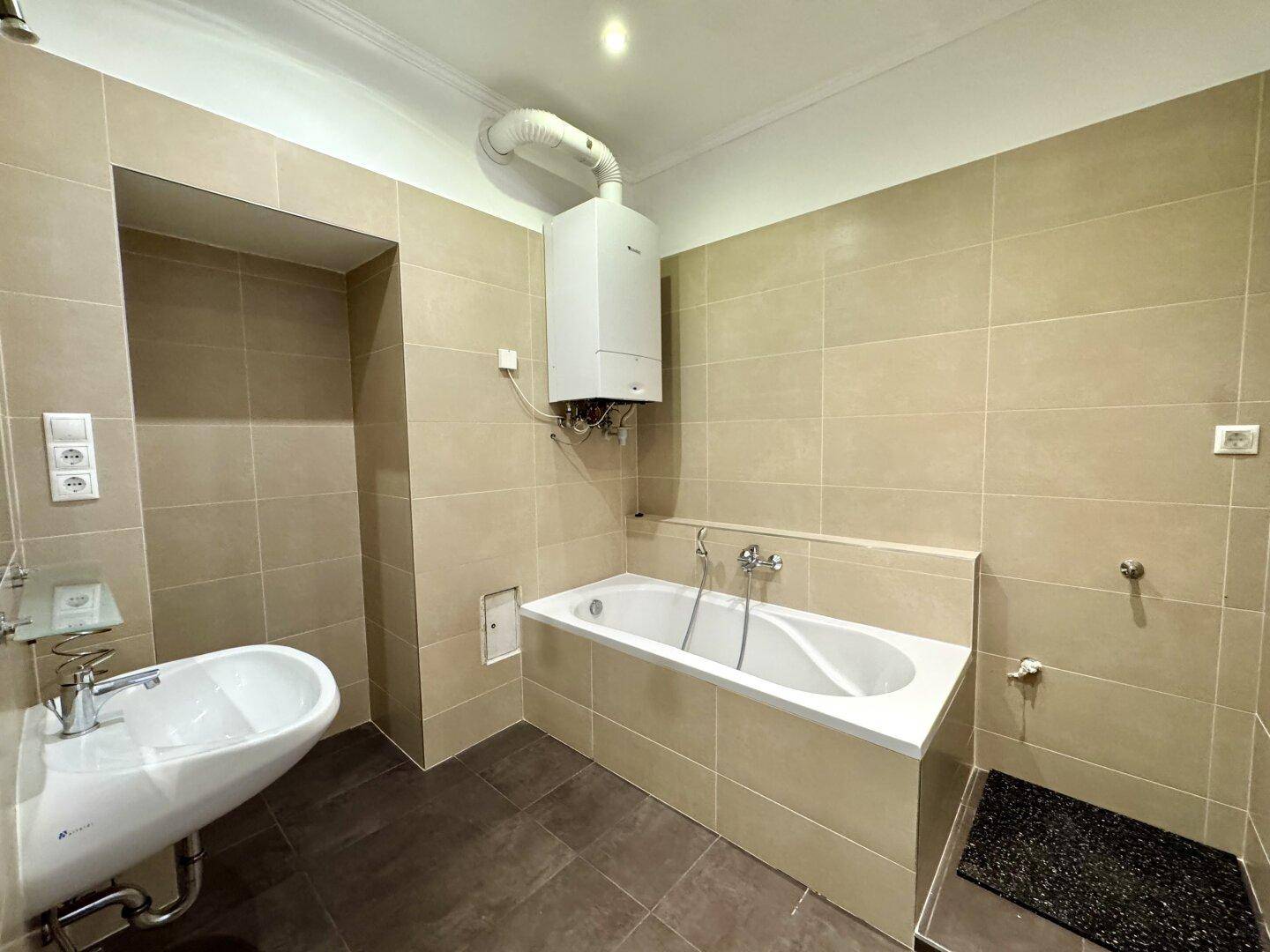 Badezimmer mit Badewanne, Waschbecken und Gastherme, gefliest in warmen Beigetönen.
