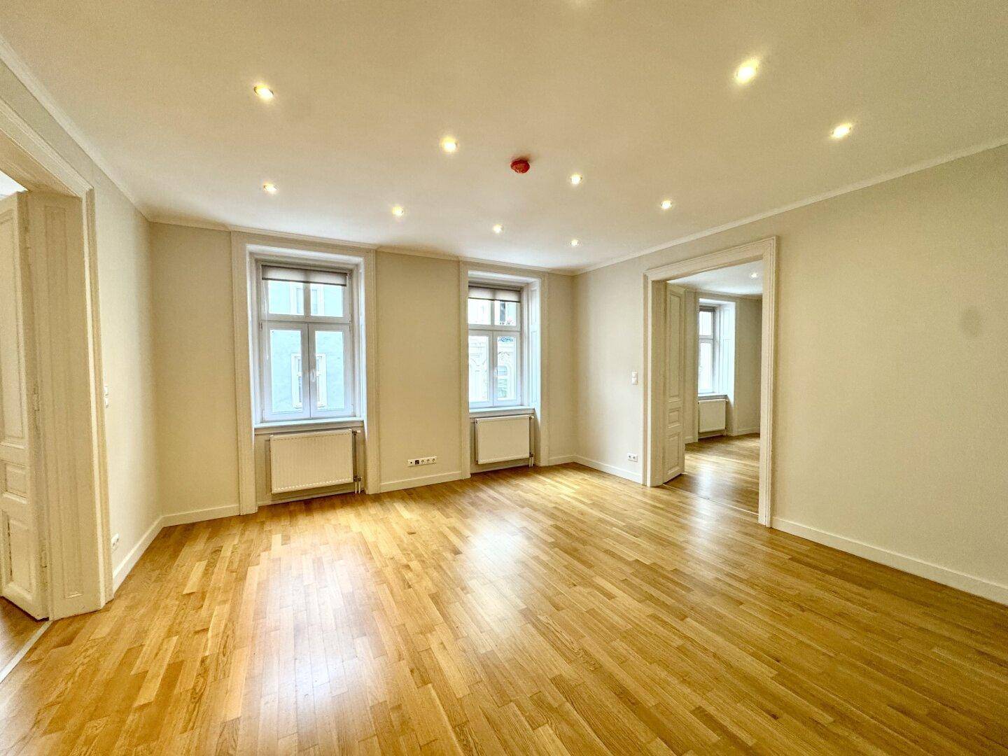 Großes Zimmer mit Parkettboden und zwei Fenstern, das viel Tageslicht hereinlässt.