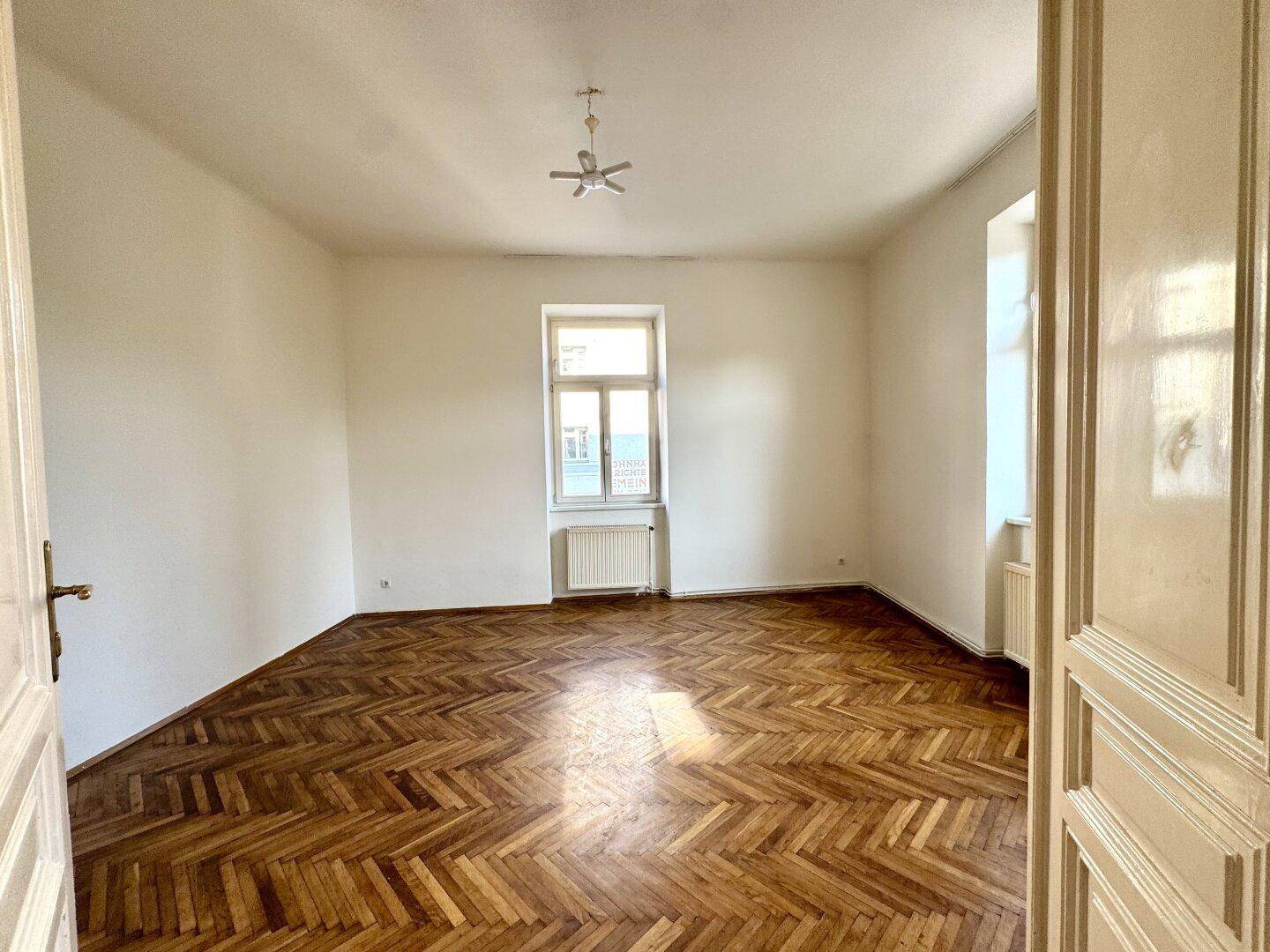 Geräumiges Zimmer mit klassischem Fischgrätparkett, hohen Decken und großem Fenster.