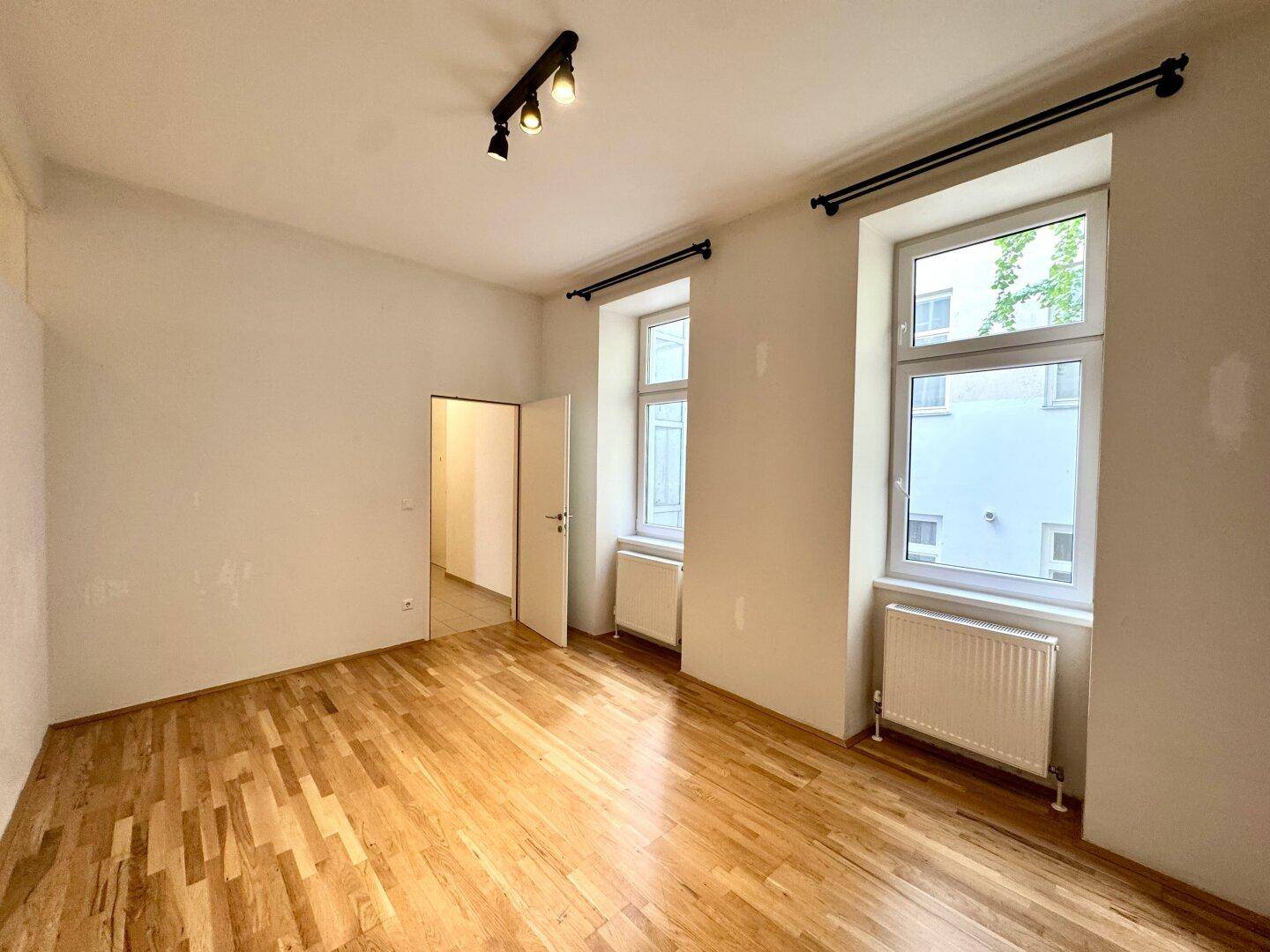 Helles Zimmer mit Parkettboden und zwei Fenstern, eines davon mit Blick auf den Innenhof.