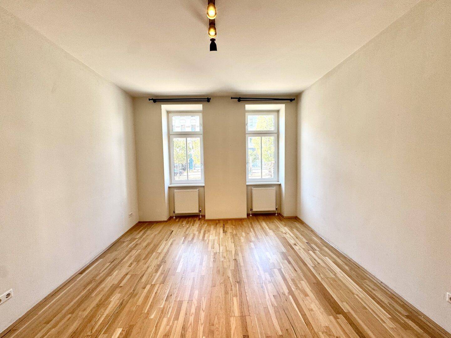 Lichtdurchflutetes Zimmer mit zwei großen Fenstern und klassischem Parkettboden, bereit zur individuellen Gestaltung.