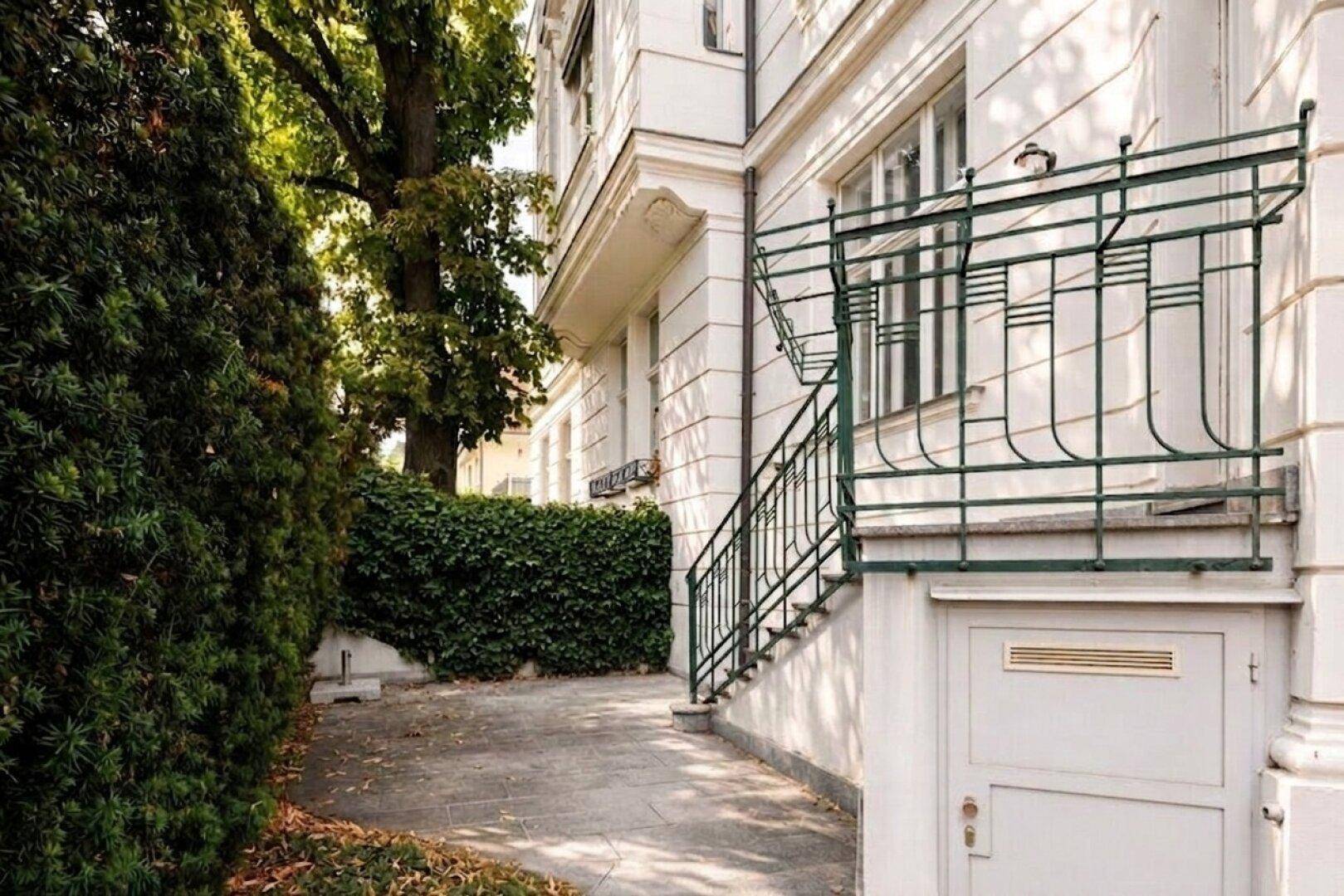 Seitenansicht eines historischen Gebäudes mit einer Außentreppe und einer hohen, dichten Hecke.