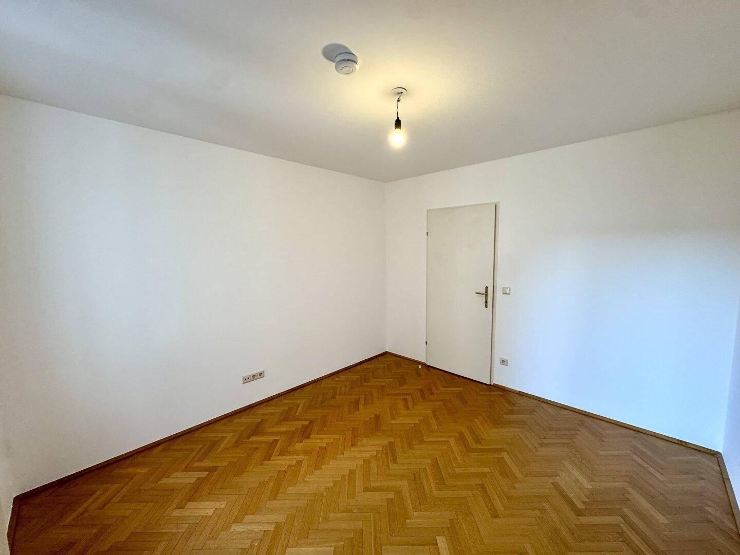 Leeres Zimmer mit Fischgrätparkett und weiß gestrichenen Wänden, bereit zur Nutzung.