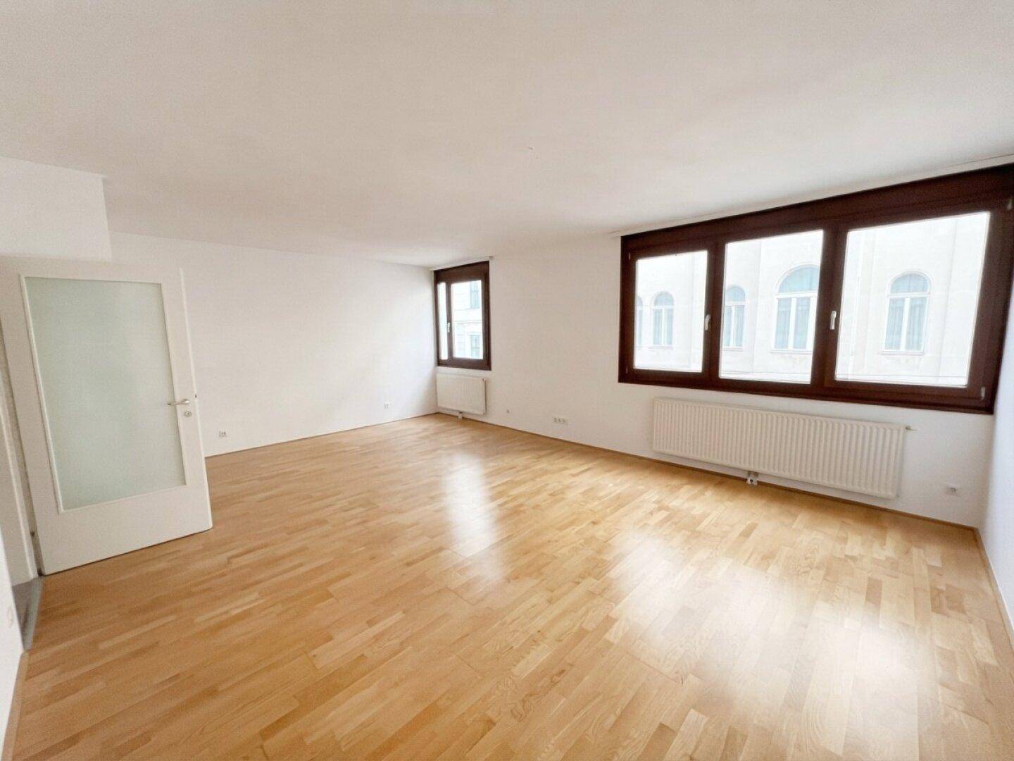 Heller, leerer Raum mit Parkettboden, großen Fenstern und viel Tageslicht.