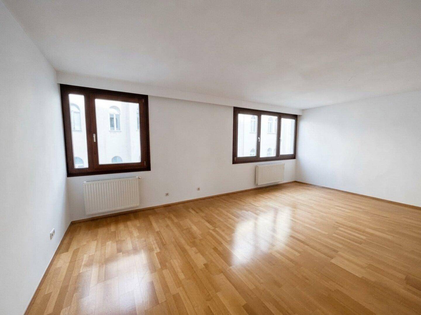 Geräumiges, leeres Zimmer mit hellem Parkettboden und zwei großen Fenstern.