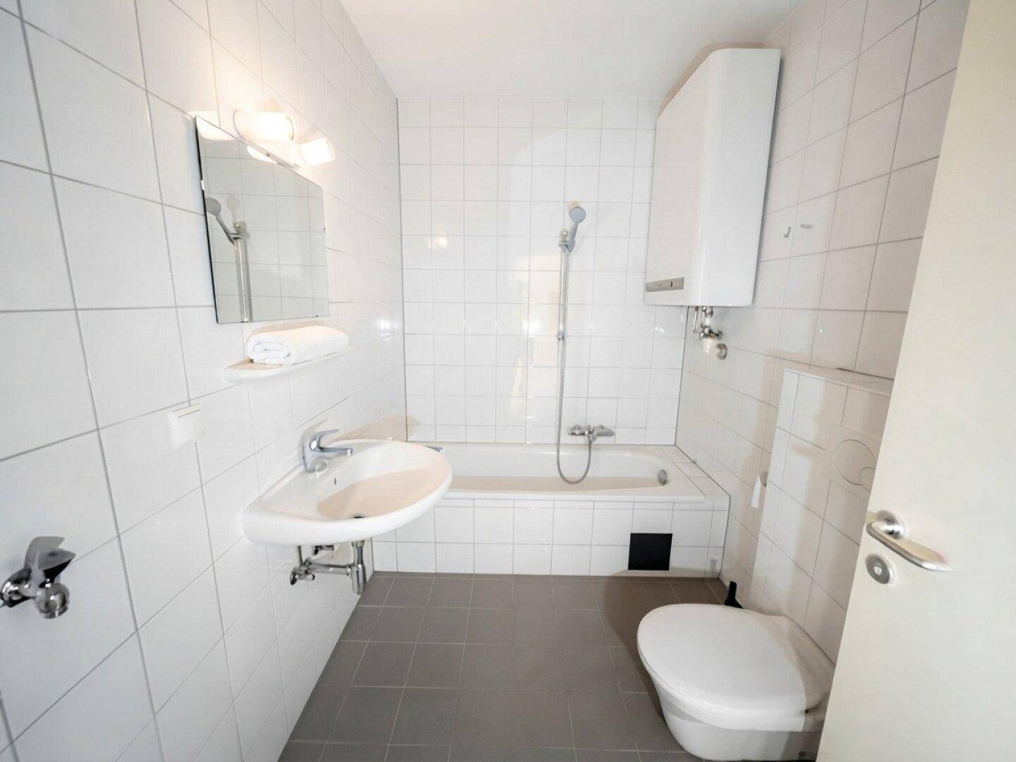 Helles Badezimmer mit weißen Fliesen, Badewanne mit Duschfunktion, Waschbecken und WC.