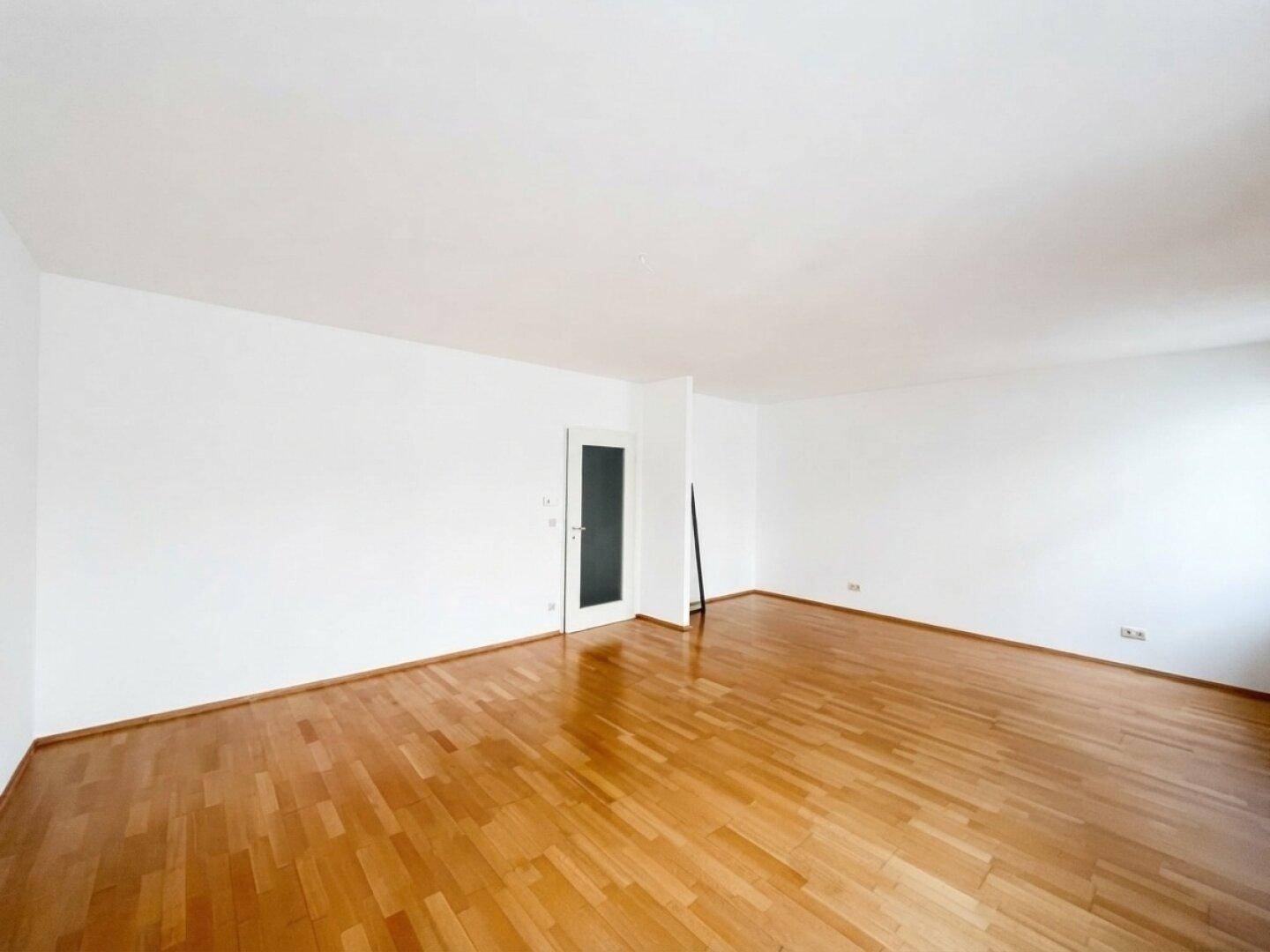 Leerer Wohnraum mit glänzendem Parkettboden und einer modernen Tür, die zu weiteren Räumen führt.