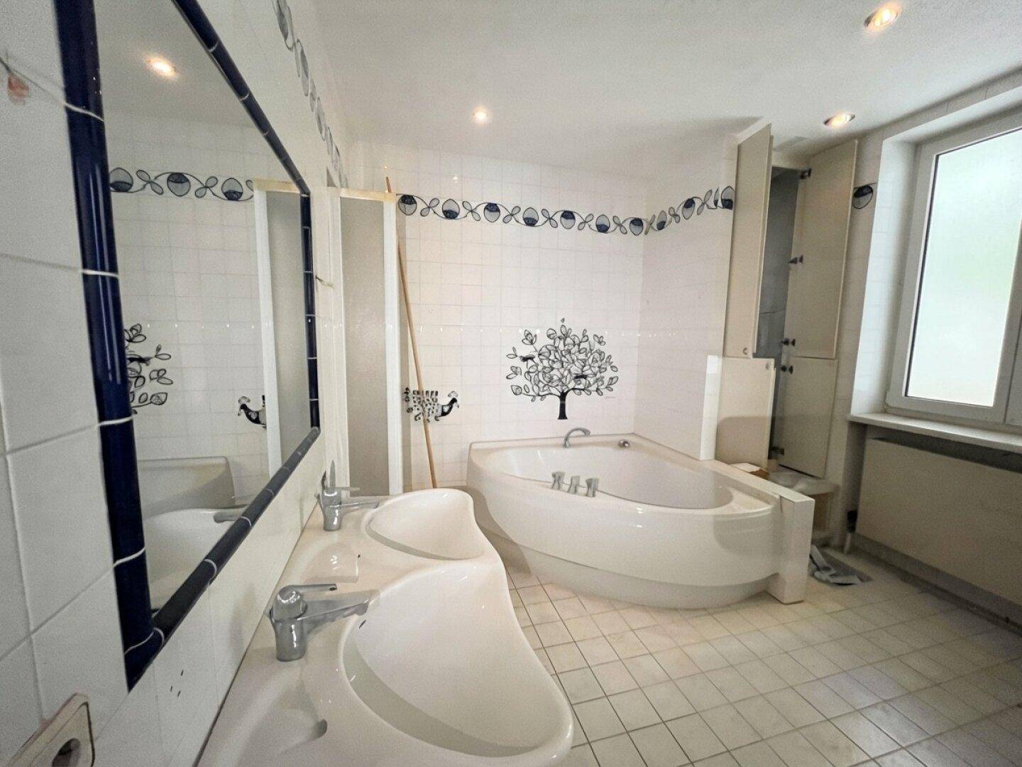 Badezimmer mit Doppelwaschbecken, Eckbadewanne und Fliesen im 80er-Jahre-Stil, renovierungsbedürftig.
