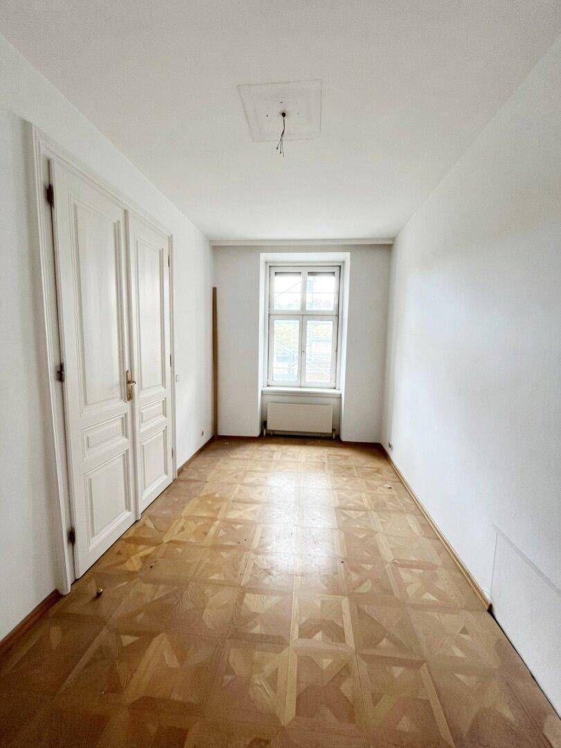 Helles Schlafzimmer in einem Altbau mit Fischgrätparkett und hohen Flügeltüren, renovierungsbedürftig.