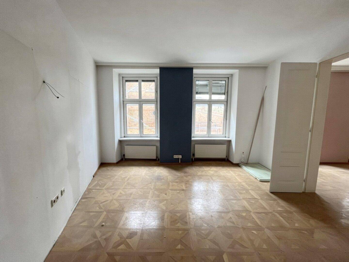 Geräumiges Wohnzimmer in einem Altbau mit Fischgrätparkett und dunkler Akzentwand, renovierungsbedürftig.