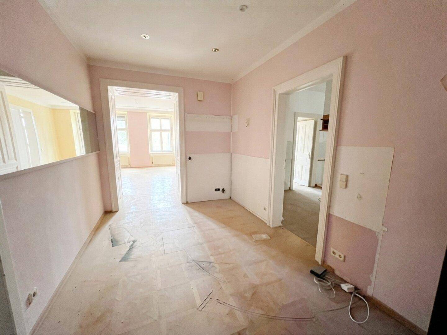 Langer Flur in einem Altbau mit rosa Wänden und Renovierungsbedarf, führt zu weiteren Räumen.