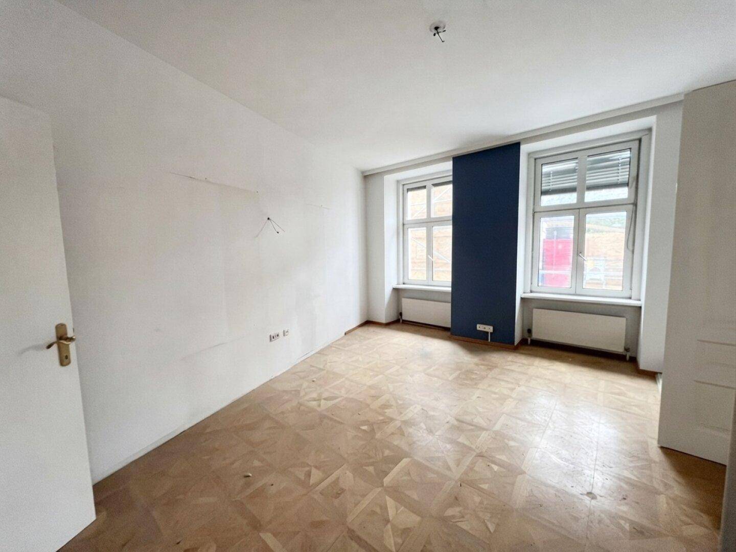 Helles Schlafzimmer in einem Altbau mit Fischgrätparkett und dunkler Akzentwand, renovierungsbedürftig.