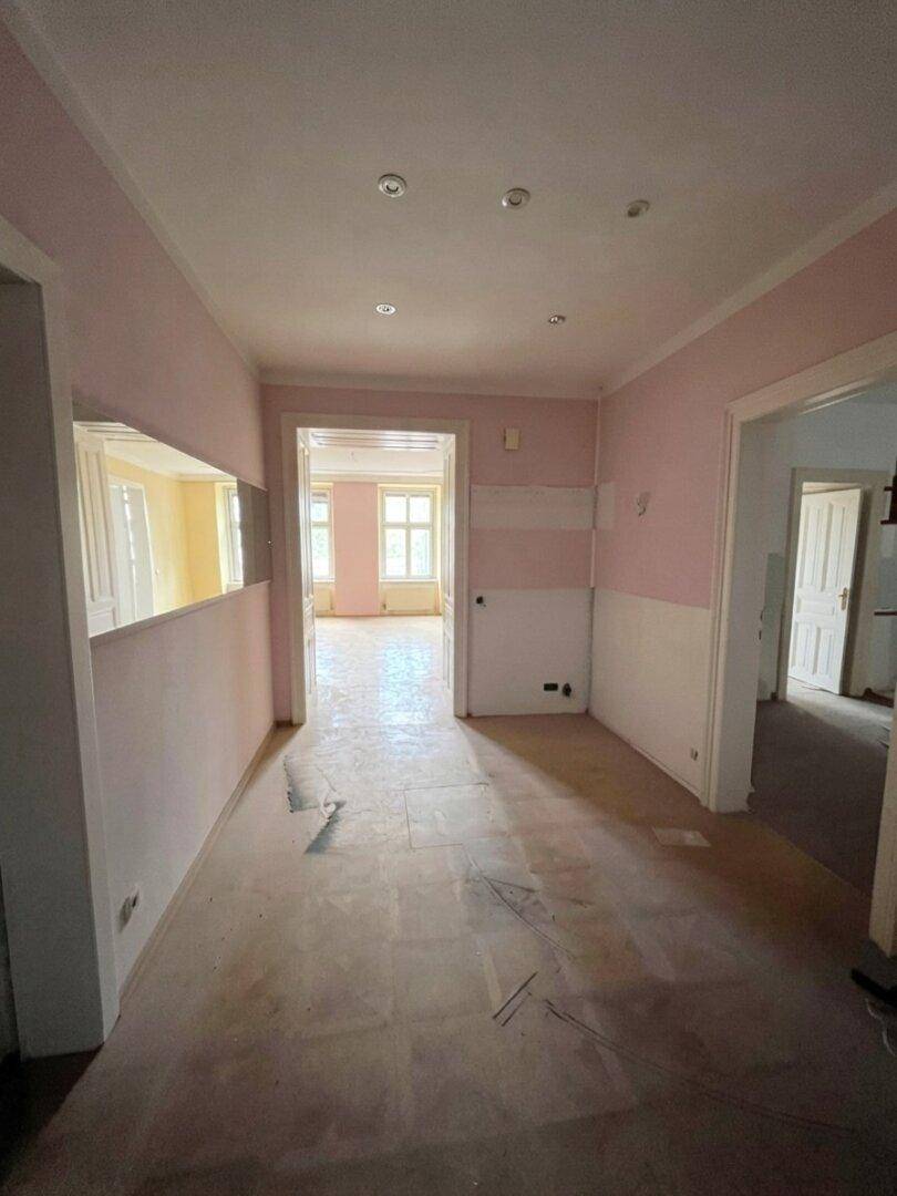 Langer Flur in einem Altbau mit rosa Wänden und Renovierungsbedarf, führt zu weiteren Räumen.