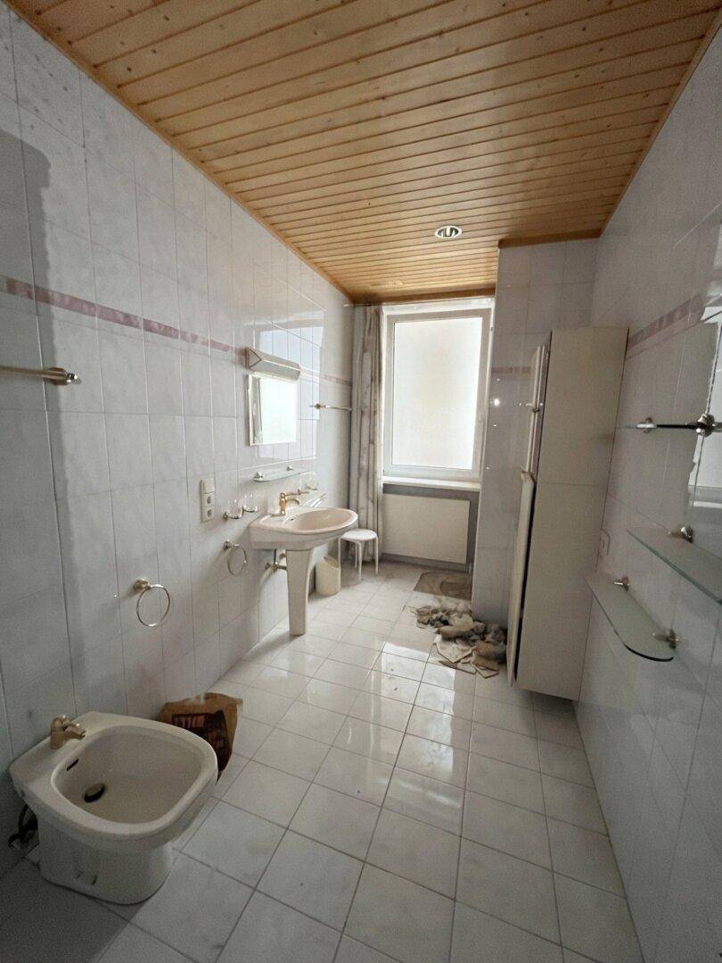Badezimmer mit weißen Fliesen, Holzdecke, Waschbecken, Bidet und einem Fenster für natürliche Belüftung.