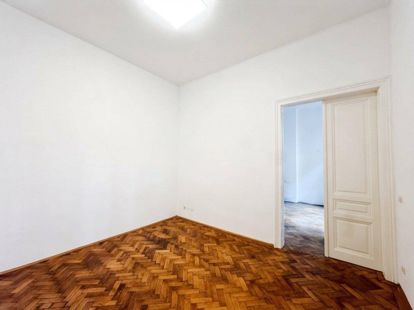 Leeres Zimmer mit Fischgrätparkett und einer Tür, die zu einem weiteren Raum führt.