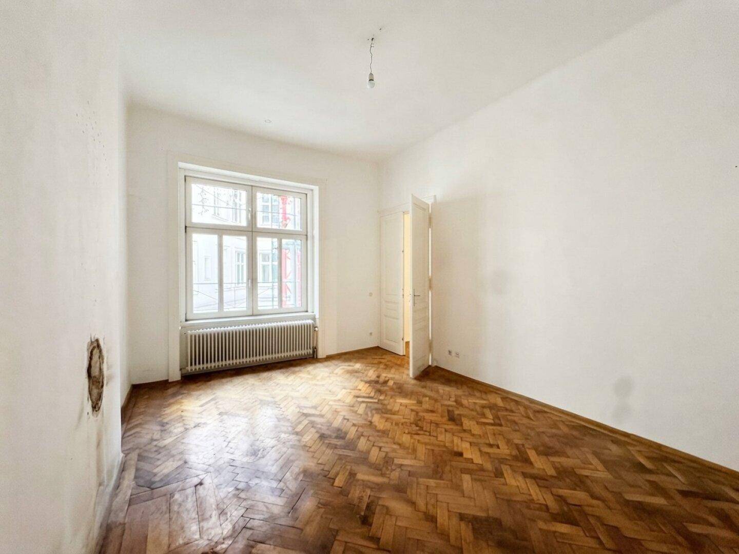 Lichtdurchfluteter Raum mit großem Fenster, Heizkörper und klassischem Fischgrätparkett.