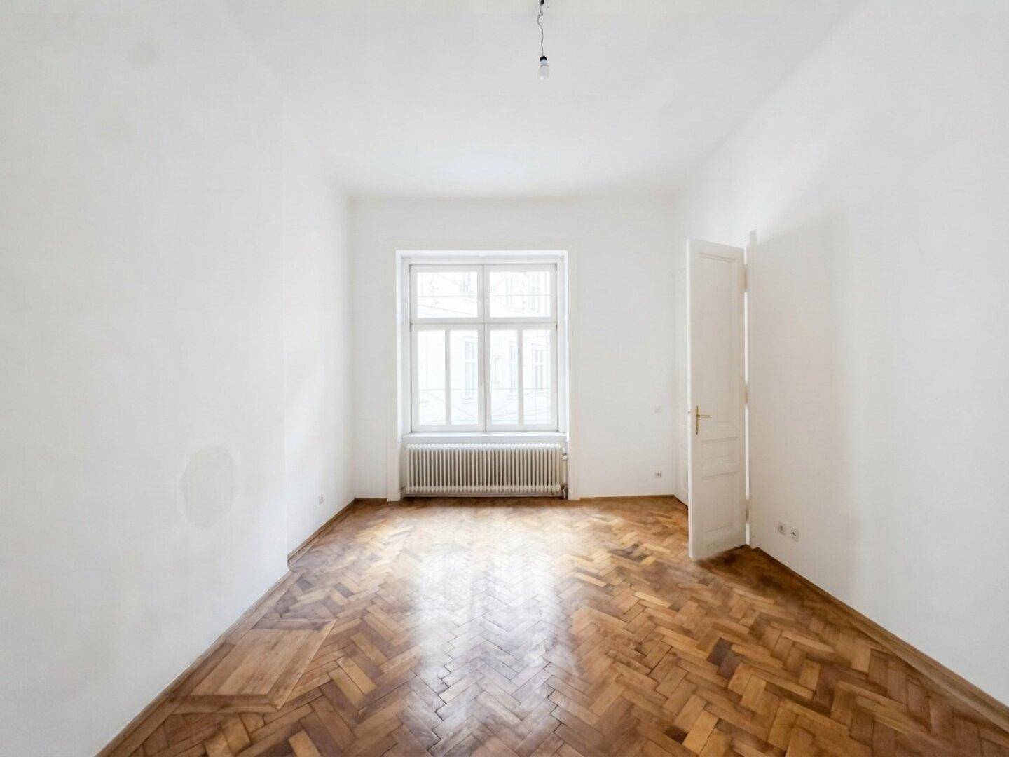 Heller, leerer Raum mit einem großen Fenster und elegantem Fischgrätparkett.