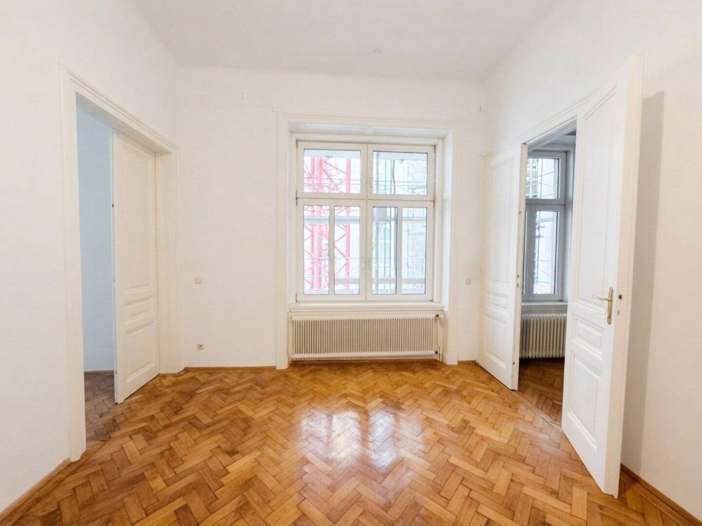 Geräumiges Zimmer mit zwei Türen, großem Fenster und klassischem Fischgrätparkett.