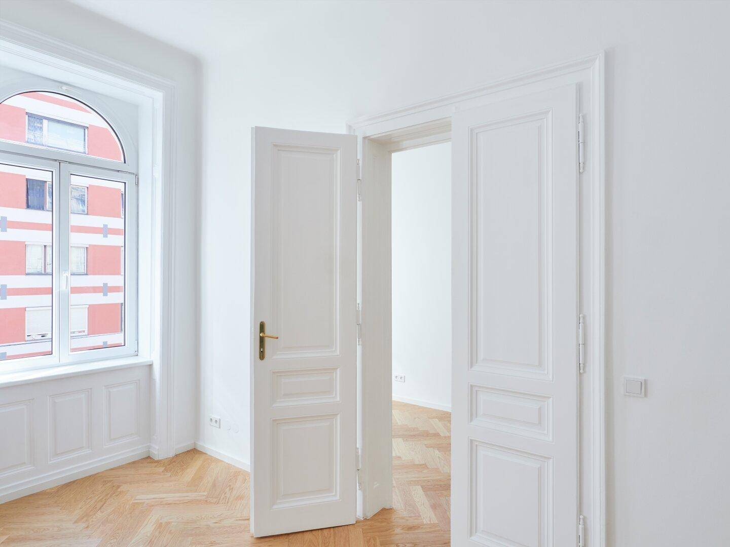 Weiße Flügeltür und hohes Bogenfenster in einem hellen Raum mit Fischgrätparkett.