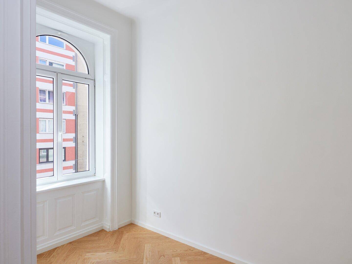 Helle Ecke eines Zimmers mit Fischgrätparkett und einem hohen Bogenfenster.