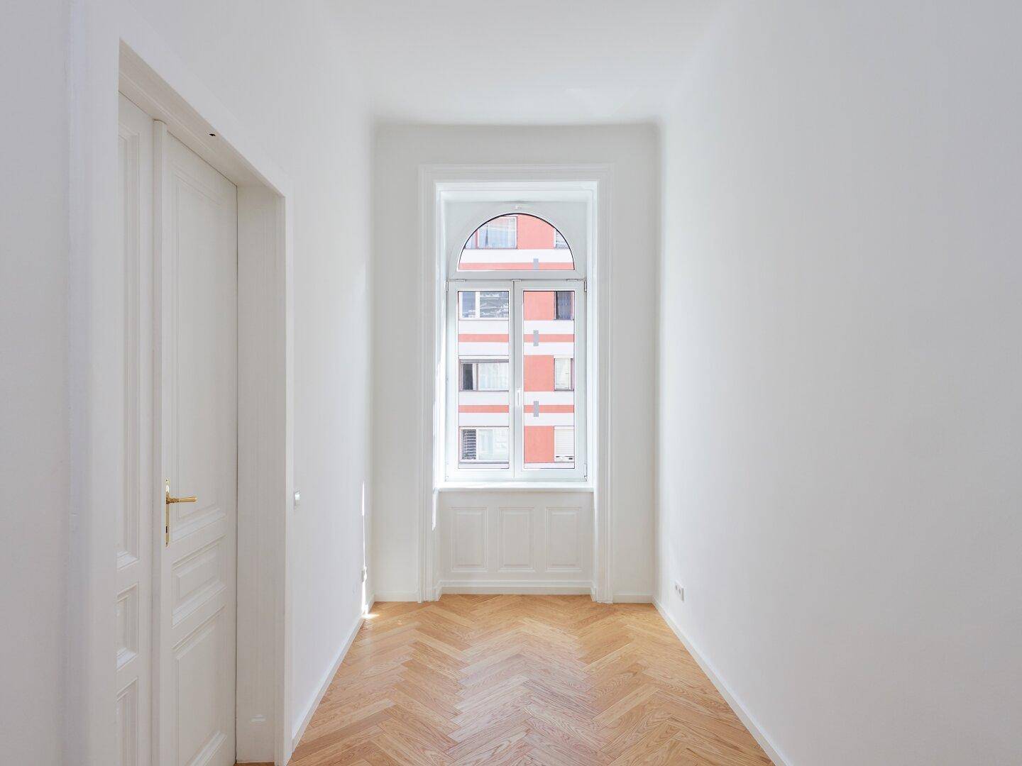 Langer, heller Flur mit Fischgrätparkett und einem Fenster am Ende des Raumes.