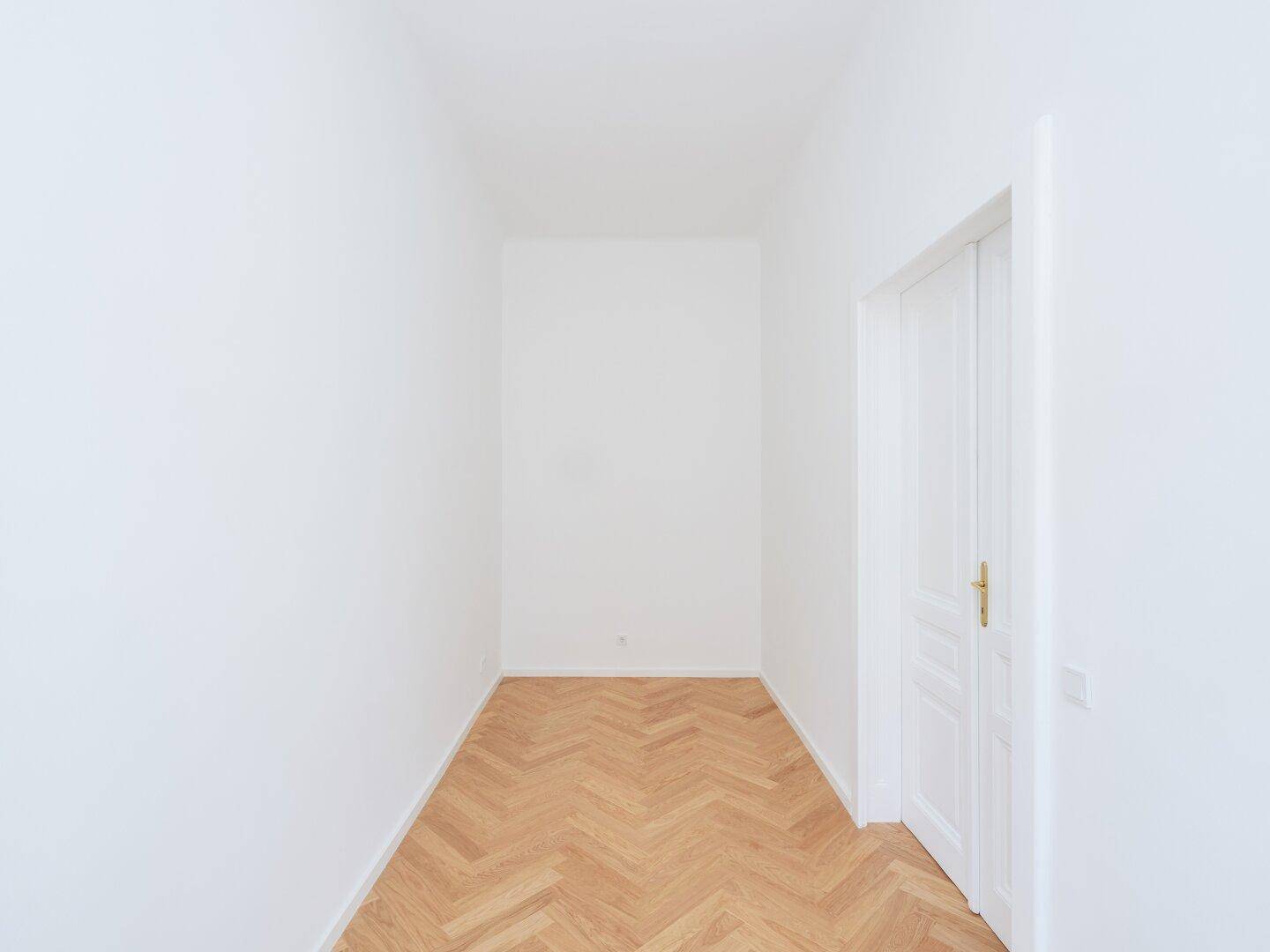 Leerer, schmaler Flur mit Fischgrätparkett und einer weißen Tür, bereit zur Nutzung.