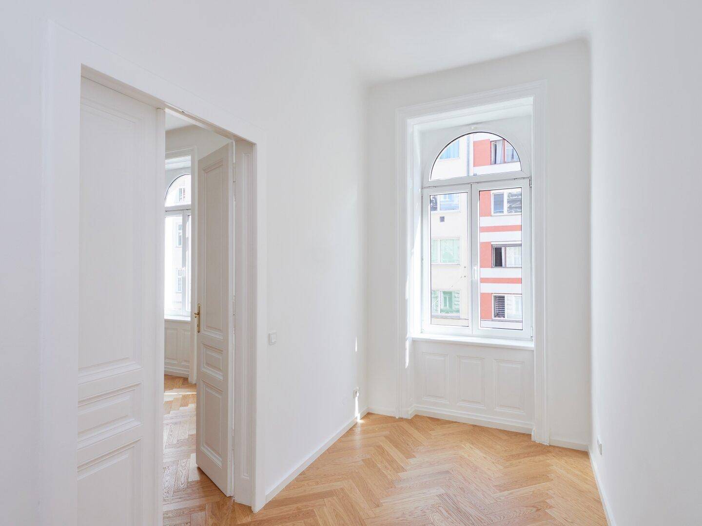 Helles Zimmer mit Fischgrätparkett und offenen Türen, die den Blick in einen weiteren Raum freigeben.