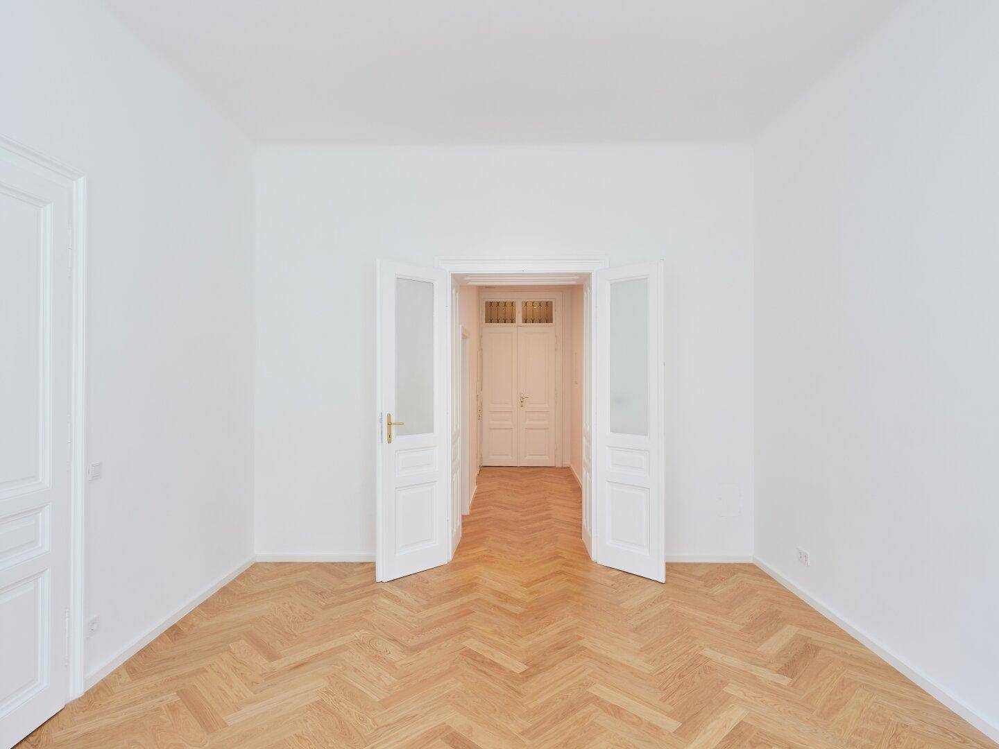 Leerer Raum mit Fischgrätparkett und eleganten Doppeltüren, die zu einem weiteren Bereich führen.