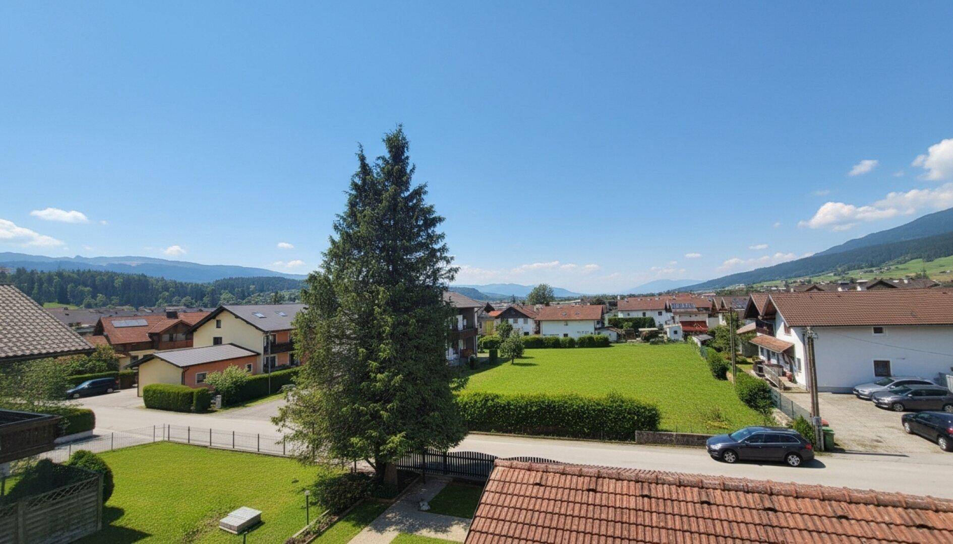 Weitläufiger Blick über eine Wohnsiedlung mit grünen Wiesen und Bergen im Hintergrund an einem sonnigen Tag.