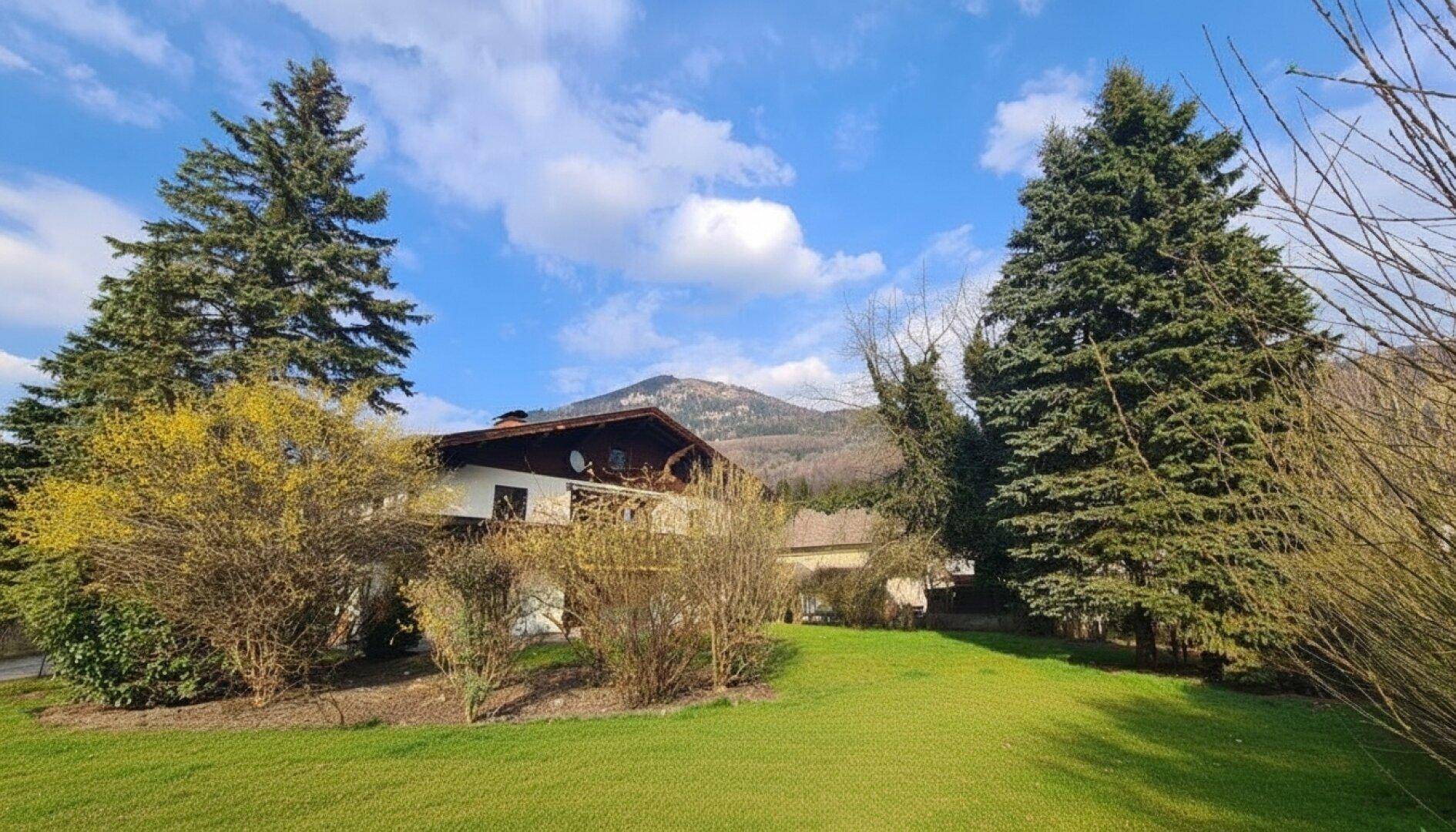 Haus mit großem Garten, umgeben von hohen Bäumen und mit weitem Blick auf die Berge unter blauem Himmel.