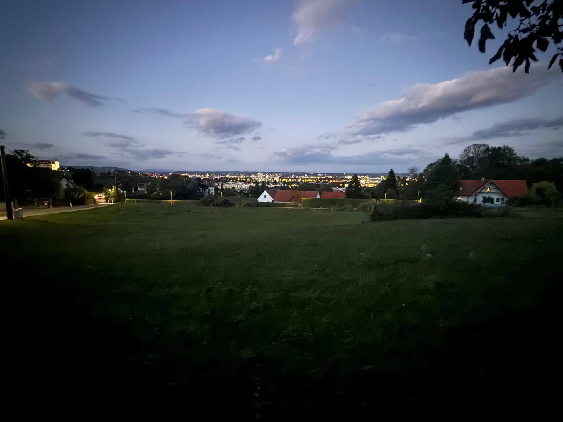 Panoramablick über ein grünes Feld auf die Lichter der Stadt unter einem bewölkten Abendhimmel mit Baumzweigen.