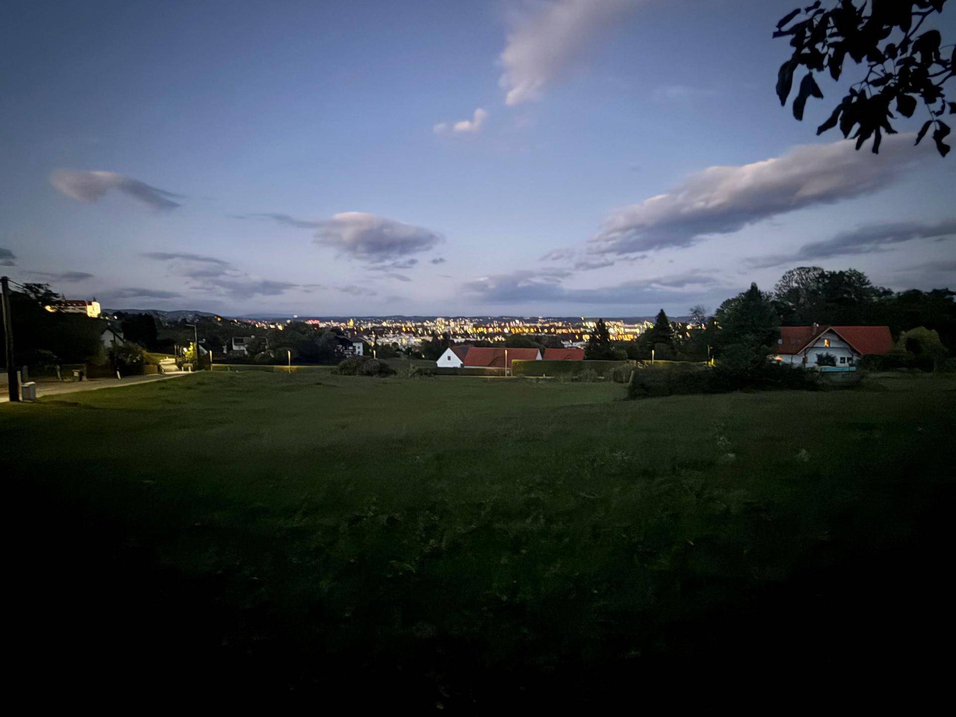 Abendlicher Panoramablick über ein grünes Feld auf die leuchtende Stadtkulisse am Horizont.