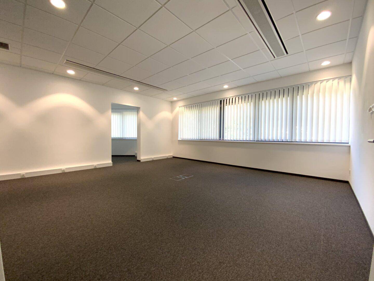 Geräumiger Büroraum mit Teppichboden, großen Fenstern mit Jalousien und guter Beleuchtung.
