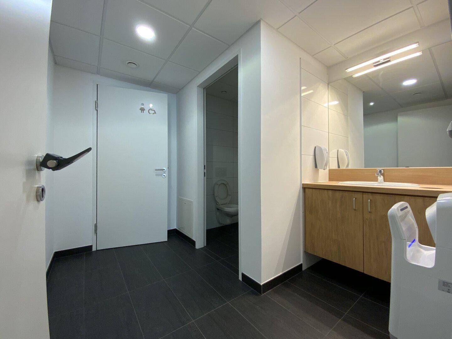 Moderner Sanitärbereich mit dunklen Fliesen, Holzwaschtisch und barrierefreiem Zugang zu den Toiletten.